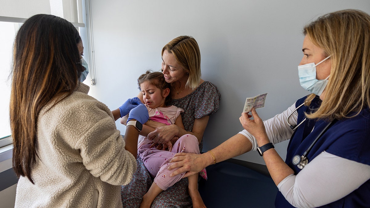 An adult woman holds an upset little girl as she is bandaged up after receiving her vaccines