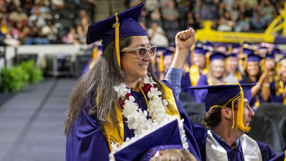A blind graduate sits in her cap and gown at commencement with her guide dog resting at her feet.