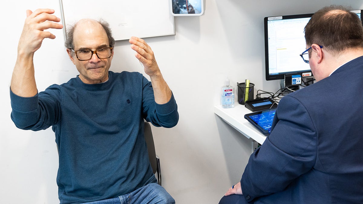 Rand Laycock, music conductor being treated for Parkinson's disease sits in an office next to a man in a suit facing away from the camera and towards a desk with a computer and tablet on it. Sat in front of a whiteboard and what appears to be a blood pressure machine in the background, Laycock dresses casually as he lifts bits hands into the air, as if he is conducting an orchestra