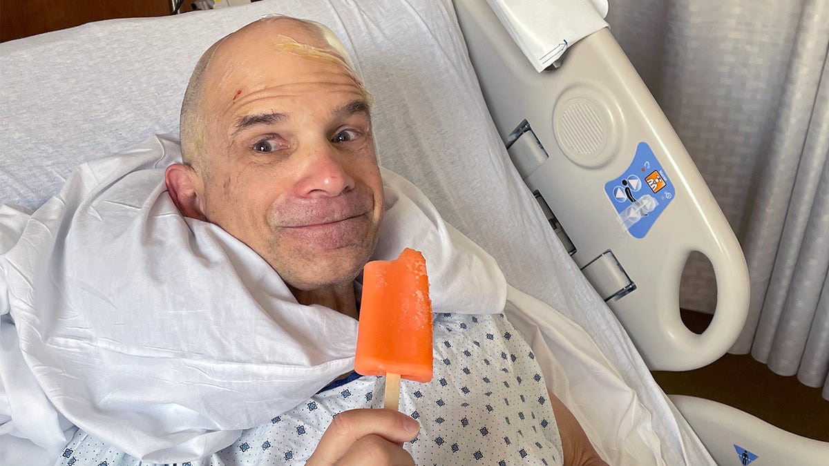 Rand Laycock sits upward in a hopsital bed wearing a gown, smiling into the camera with eyes wide as he holds an orange popsicle thay has been bitten into