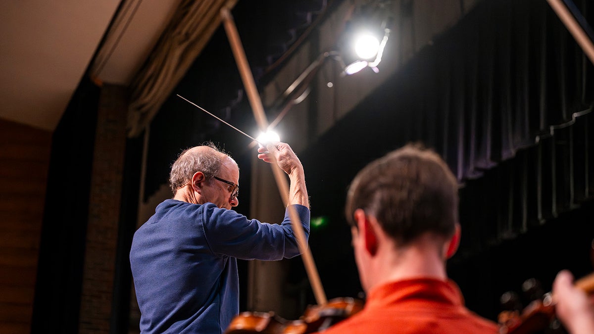 Rand Laycock pictured from behind as he leads an orchestra, wand in hand. Another person is pictured in the foreground.