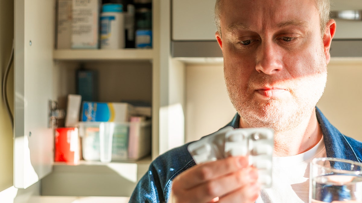 Man staring at a painkiller packet in his kitchen while holding a glass of water.