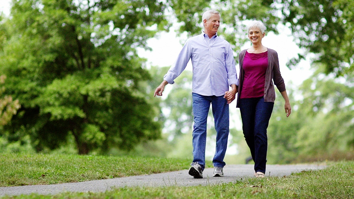 Senior couple walking in park on path with trees in the background.