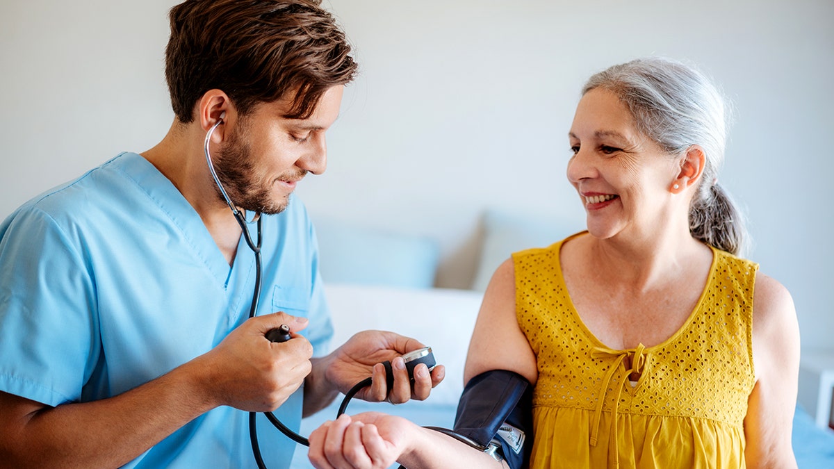 A nurse in blue scrubs checks the blood pressure of a smiling woman.