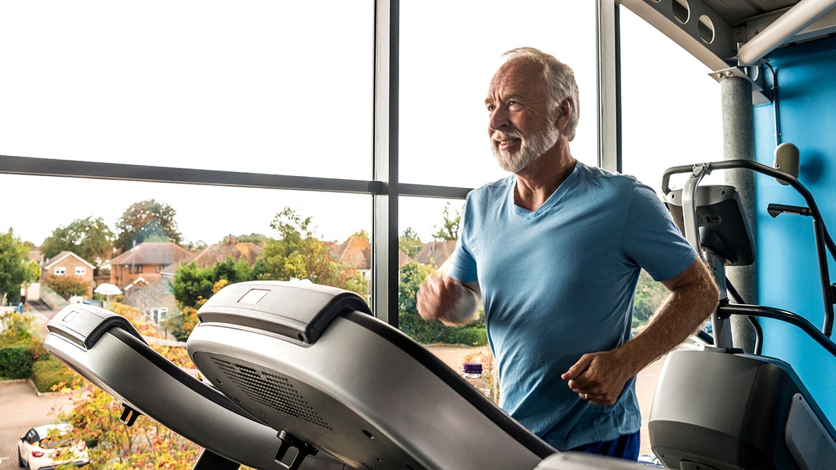 Older man running on a treadmill while smiling. Large window behind him.