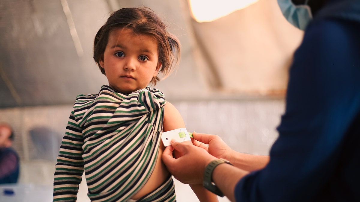 A doctor examines child's malnutrition inside a refugee camp. Malnutrition was measured using a mid-upper arm circumference belt as little girl wearing striped shirt looks into camera.