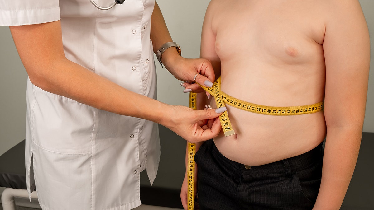 Overweight boy shirtless at doctors office as doctor measures waist circumference with measuring tape.