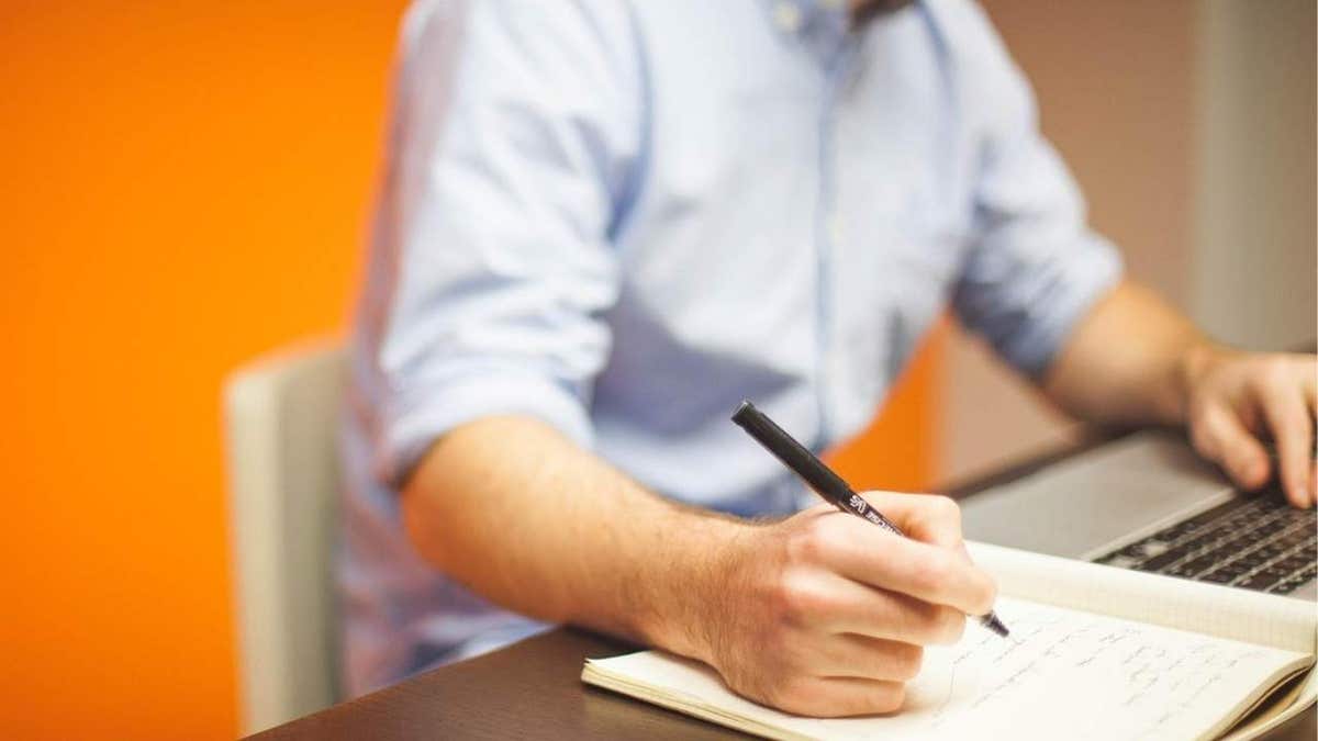 man writing in journal next to laptop while sittign at table