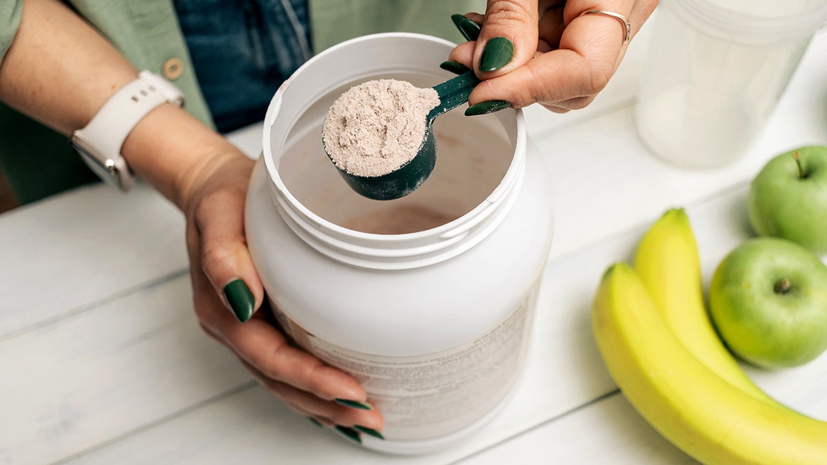 Woman in jeans and shirt holding measuring spoon with portion whey protein powder above plastic jar on white wooden table