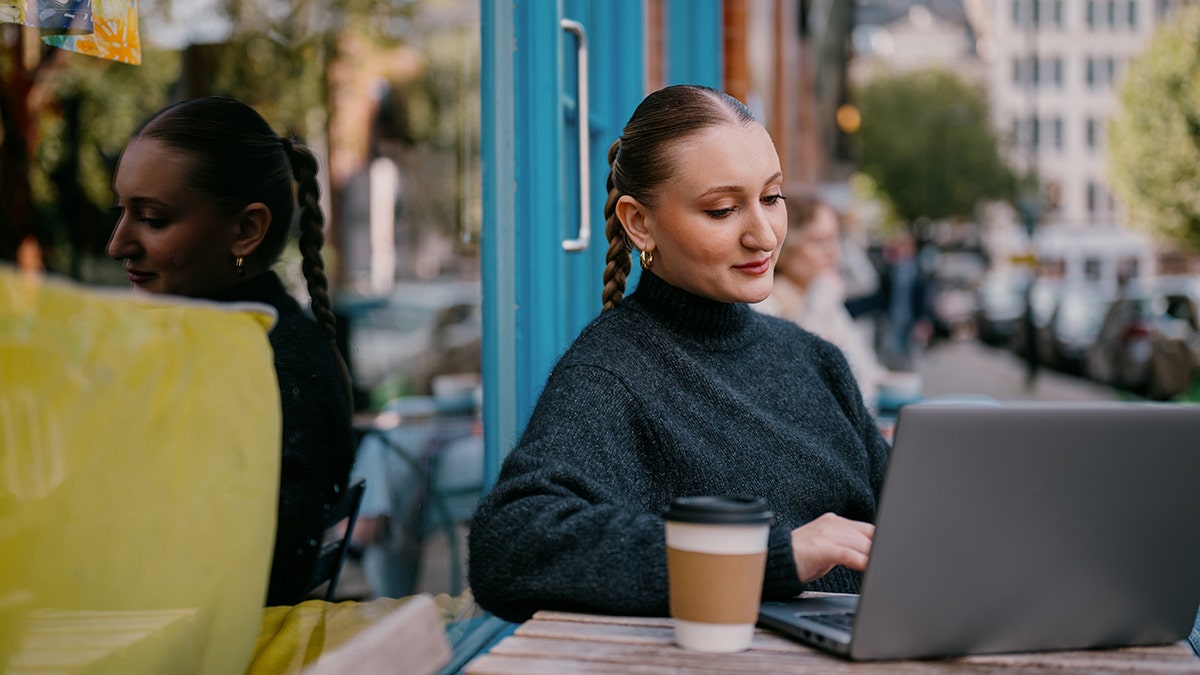 A young woman sits outside of a small, cosy coffee shop and uses her laptop while enjoying a coffee.