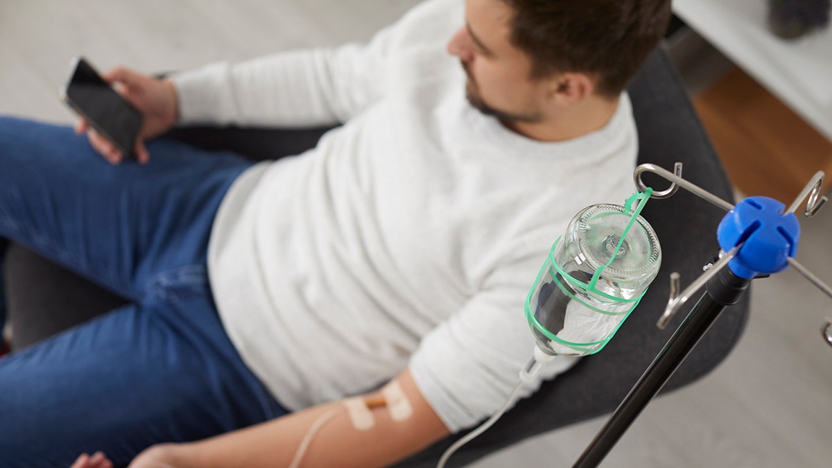 Top view photo of a man sitting in the chair and using the smartphone while receiving IV drip infusion and vitamin therapy in his blood. Person receiving injection therapy. Healthcare concept: used for piece about ketamine therapy