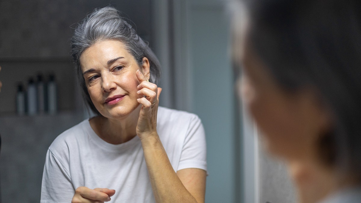 Older woman with gray hair looking in the mirror examining her cheek