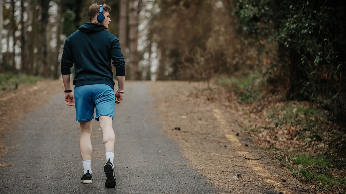 A man with headphones on walking backwards on a path in the woods while looking over his shoulder.