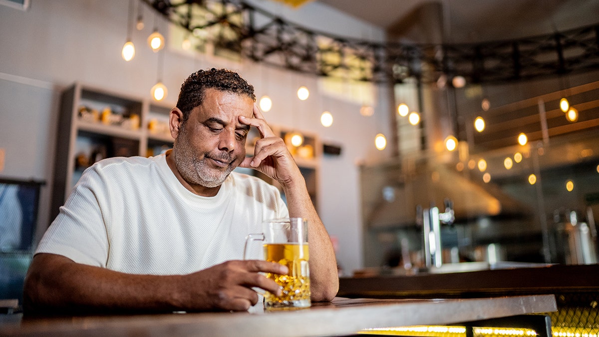 Man looking sad with head in hands, looking at bug of beer while sitting in bar, decorative lights hanging behind him.