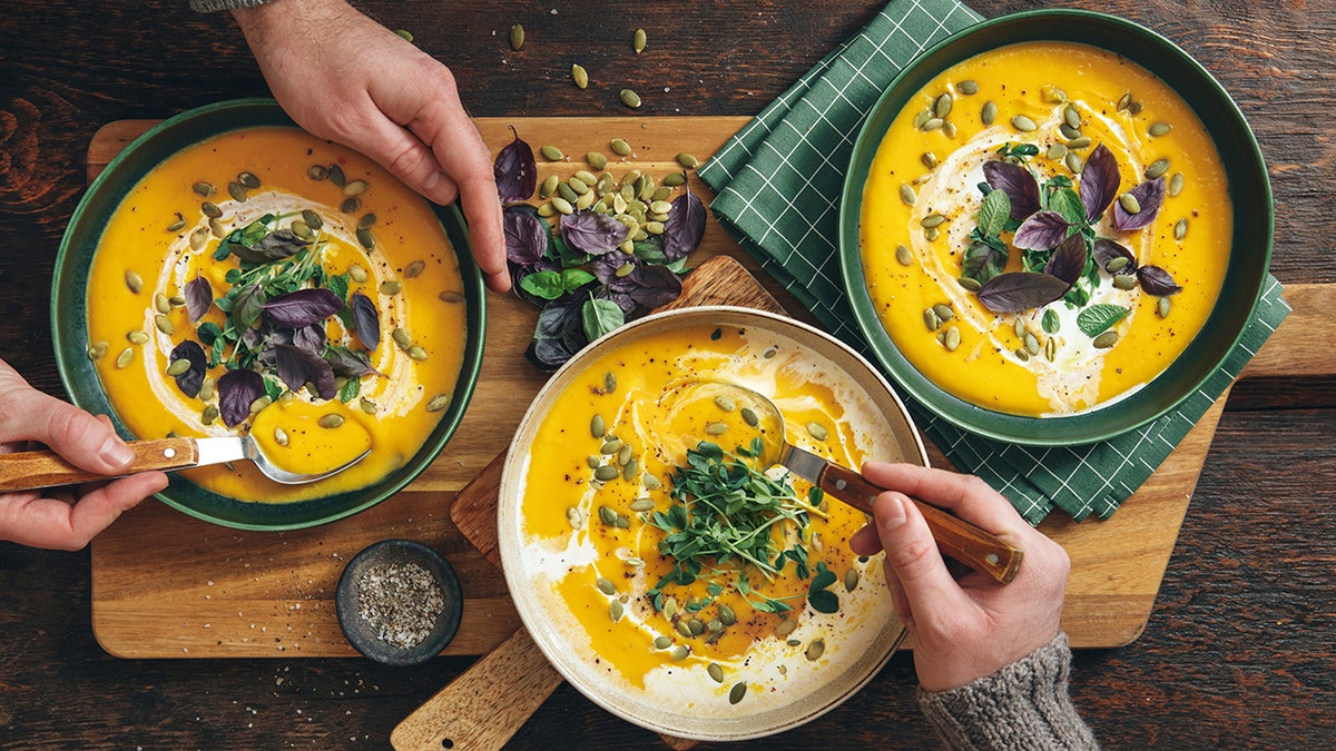 three bowls of creamy pumpkin soup with hands holding spoons