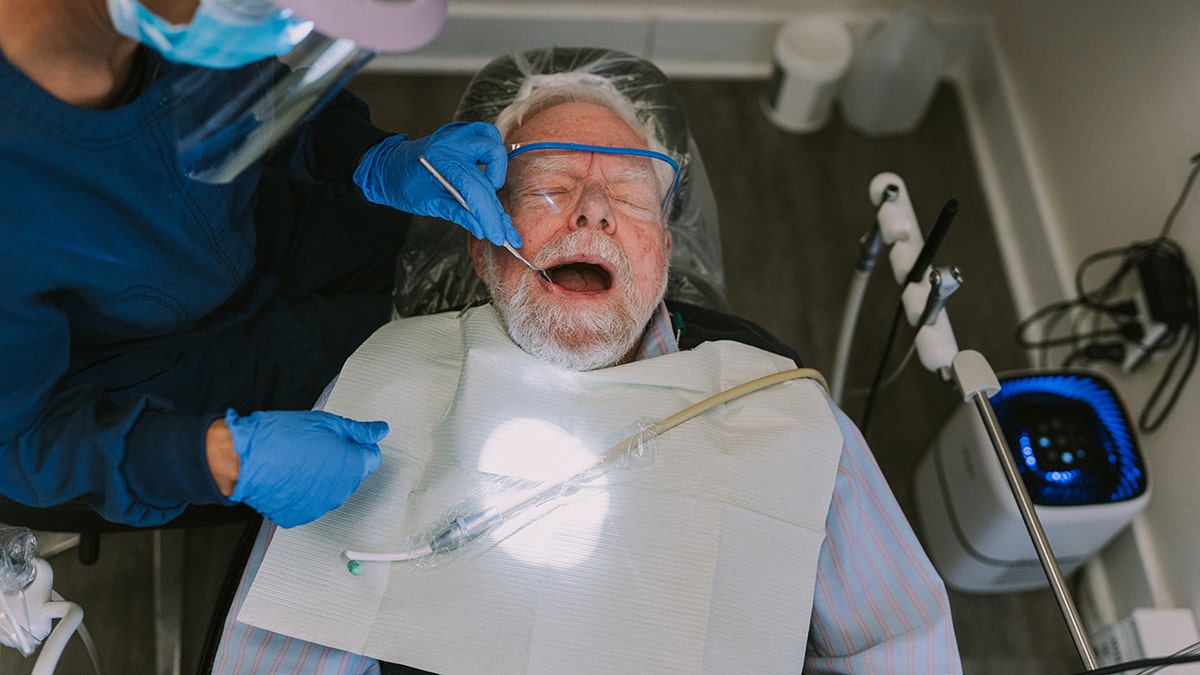 Overhead view of senior man lying in a dental chair with his mouth open as his female dentist cleans his teeth.