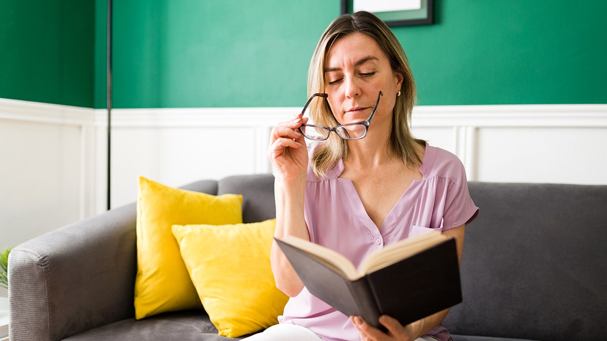 Woman peers over her reading glasses to look into book she holds in her hands