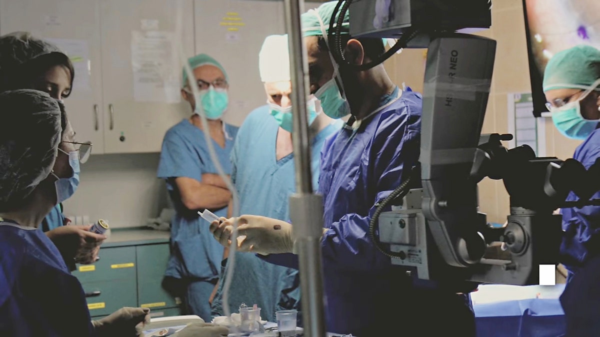 Medical staff in surgical scrubs gather around equipment during a procedure in an operating room.