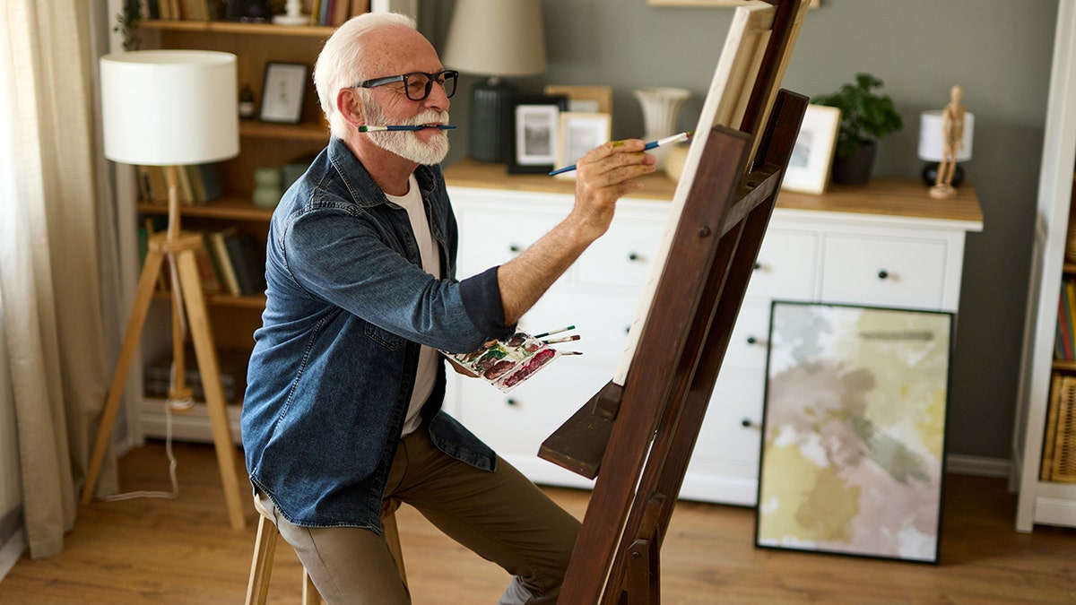Elderly man painting on a canvas at home, seen with one paintbrush between his teeth and another working as he holds paint palette, other artwork framed behind him.