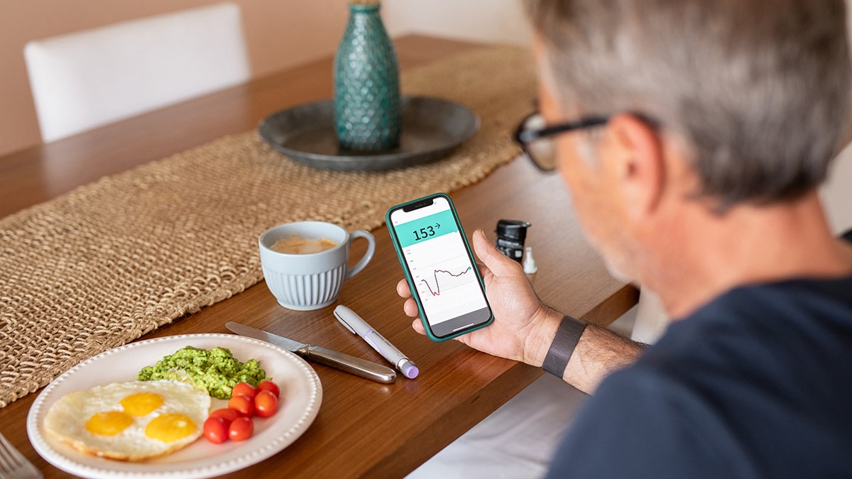 man checks blood sugar levels on phone while eating eggs, vegetables for breakfast