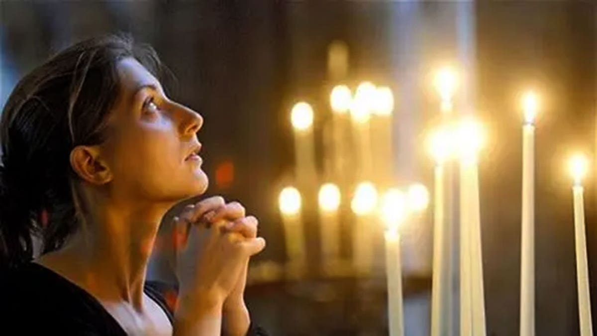 A woman kneels and looks up to heaven with candles in background.