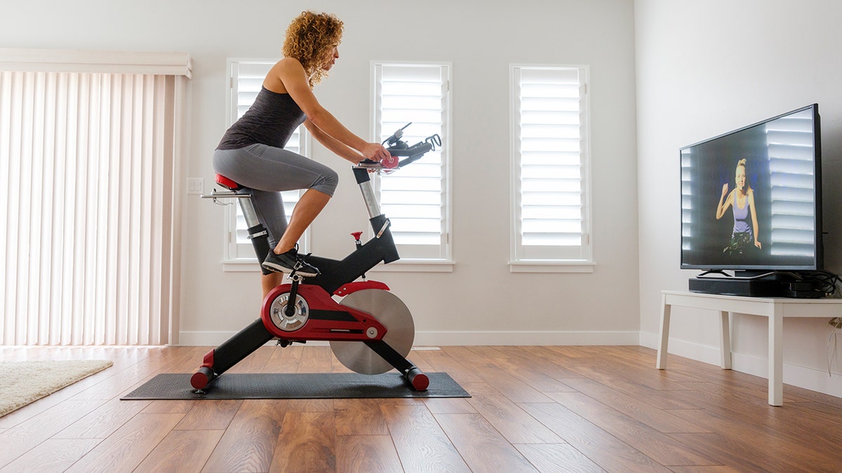 A woman is biking on a stationary bike in her home, while she watches the instructor on the television.