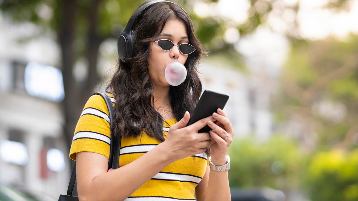 Young woman blowing bubble with gum as she looks at phone, seen outside wearing sunglasses and headphones.