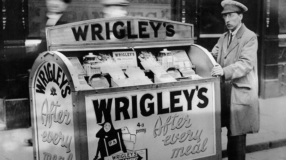 Vintage black and white image of a delivery man wearing a duster coat and a peaked cap with a Wrigley's 'chewing sweet' delivery cart, in London, England, circa 1920, walking down the street with passersby rushing past.