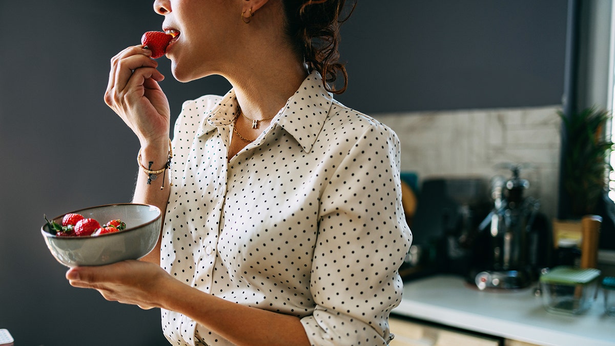 Woman in polka dot blouse eating strawberries from bowl of berries in kitchen.
