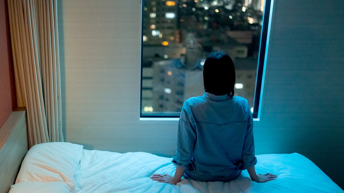 woman sits on bed and looks out window to city
