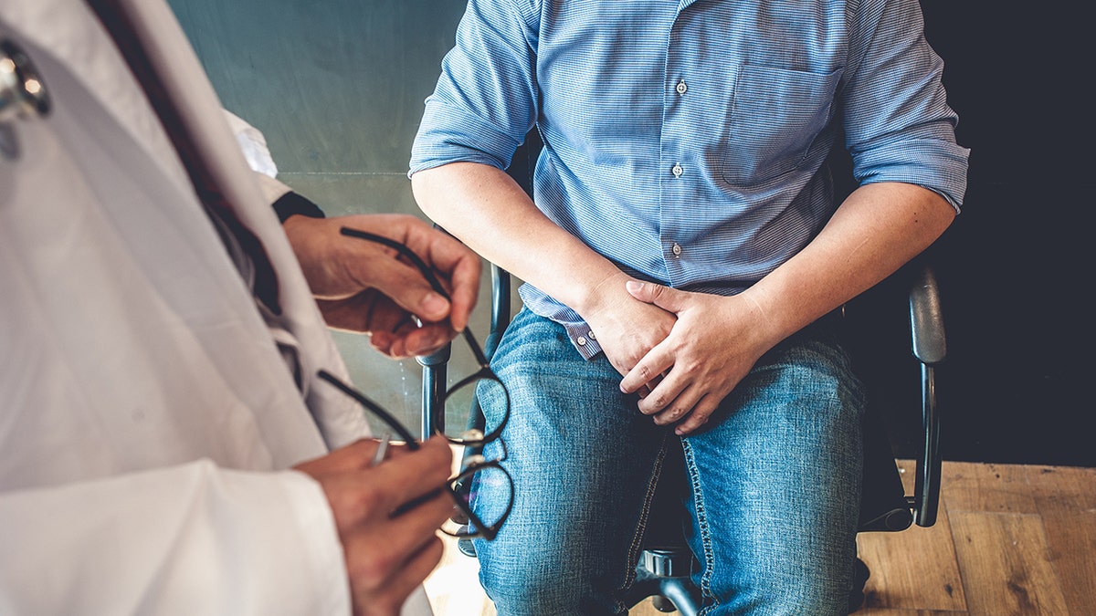 doctor holding glasses stands above man with hands over lap