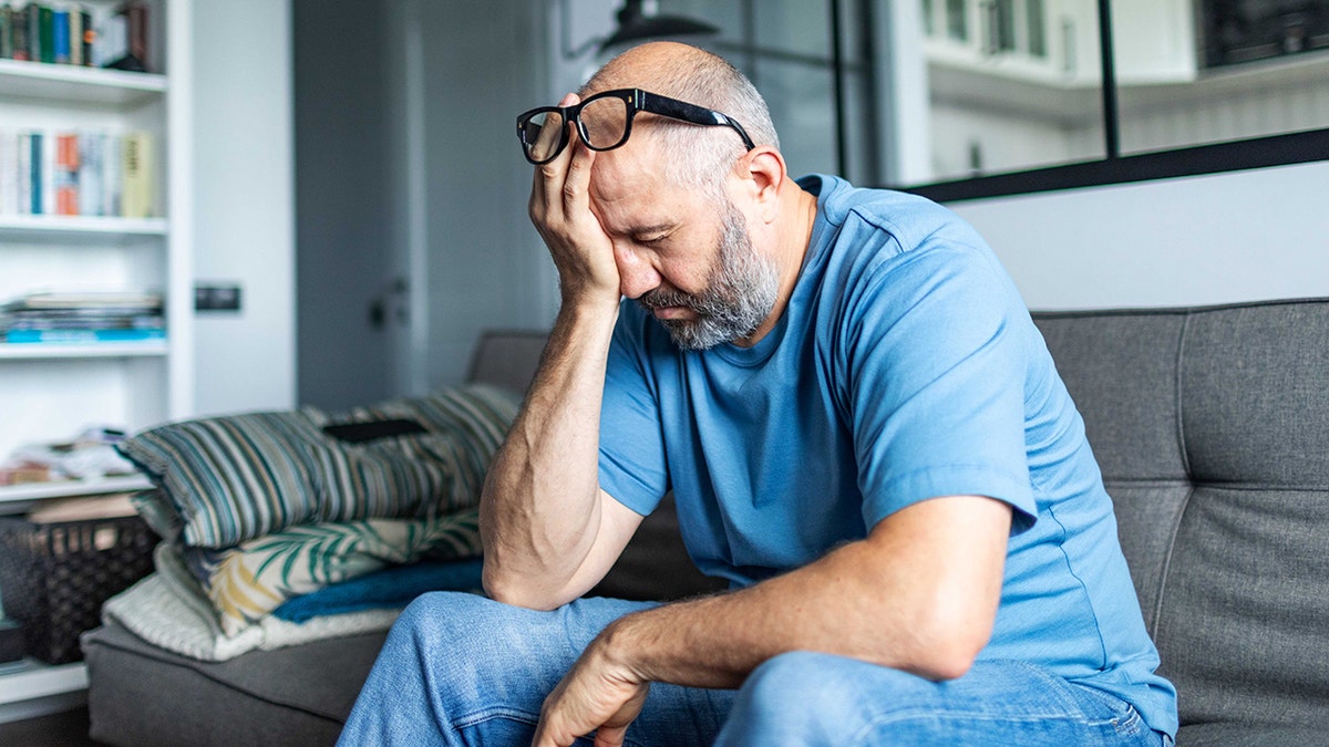 exhausted man rubs his eyes sitting on couch at home