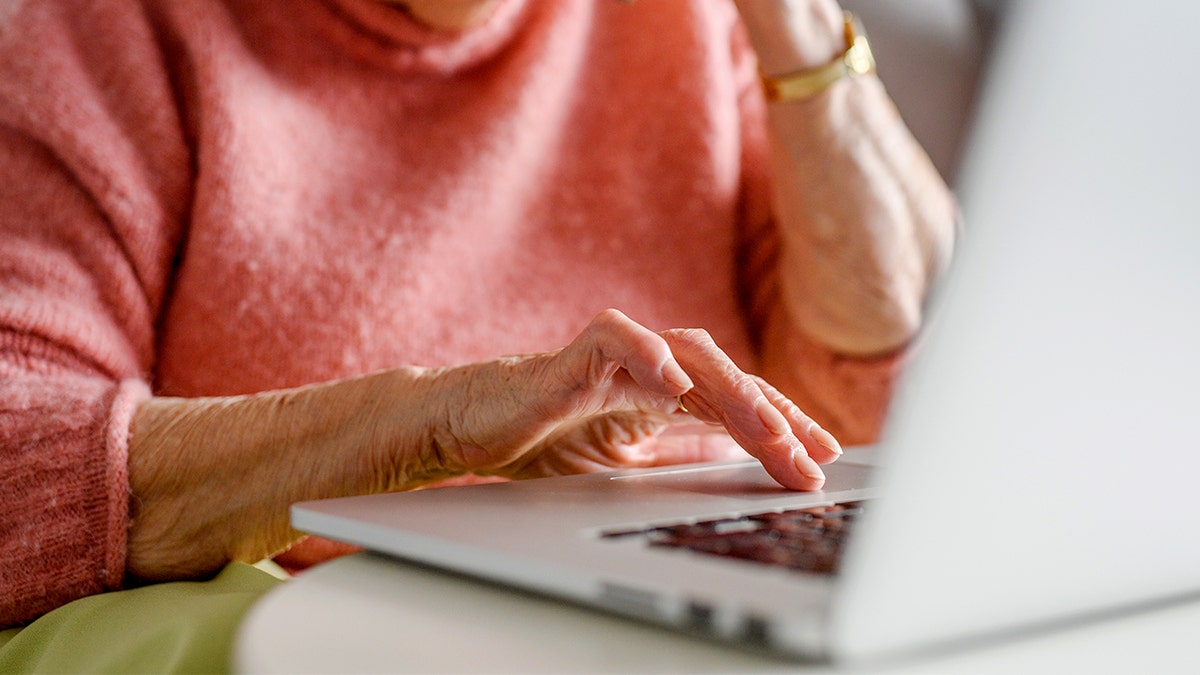 An elderly woman typing on a laptop while holding a phone.