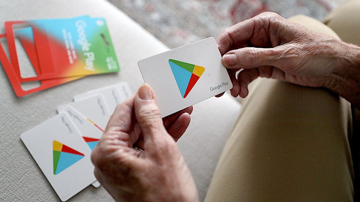 An elderly woman holds Google Play gift cards on a couch.
